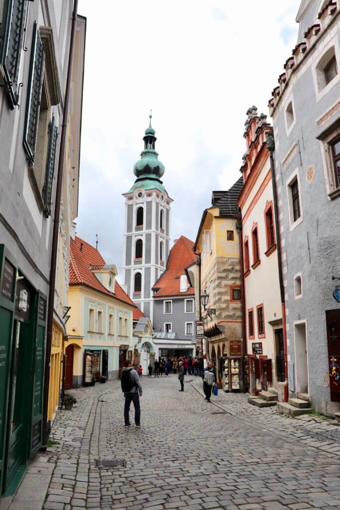 M looking up at colorful European buildings with a green-domed church beyond