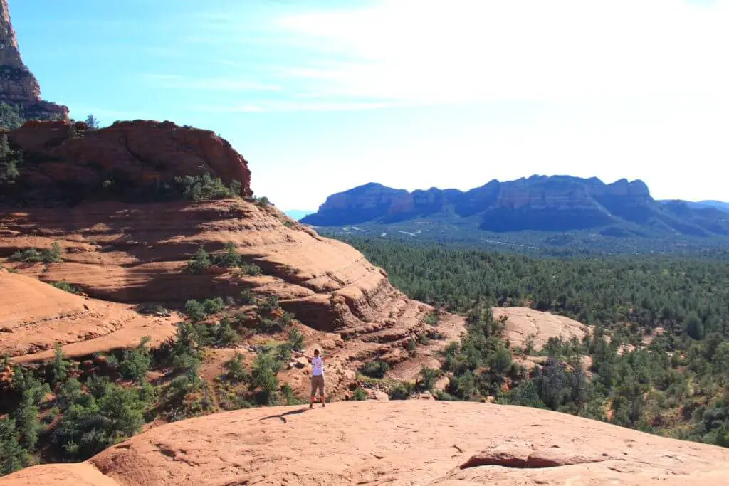 Gwen with arms spread wide in front of red rock formations and pine forest in the Southwest