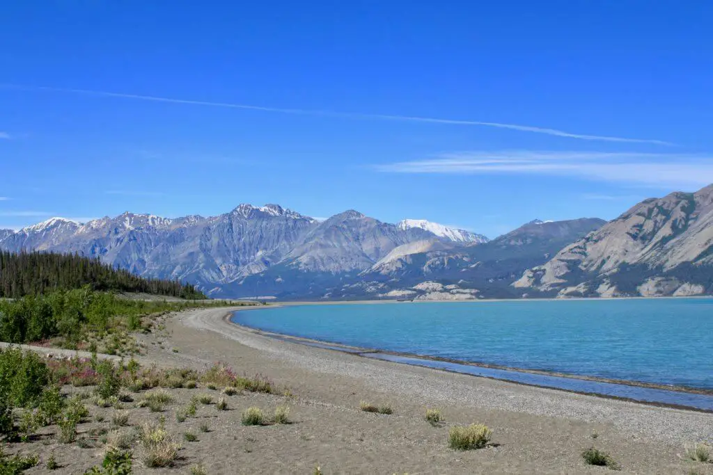 Lakeshore with bright blue water and mountains beyond. Kluane National Park is one of the best places to travel in the Yukon.