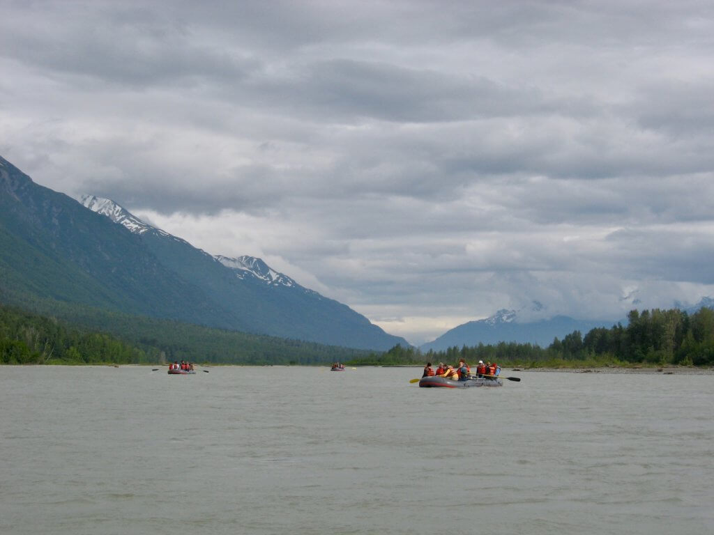 Three rafts with rafters floating down a river with mountains behind