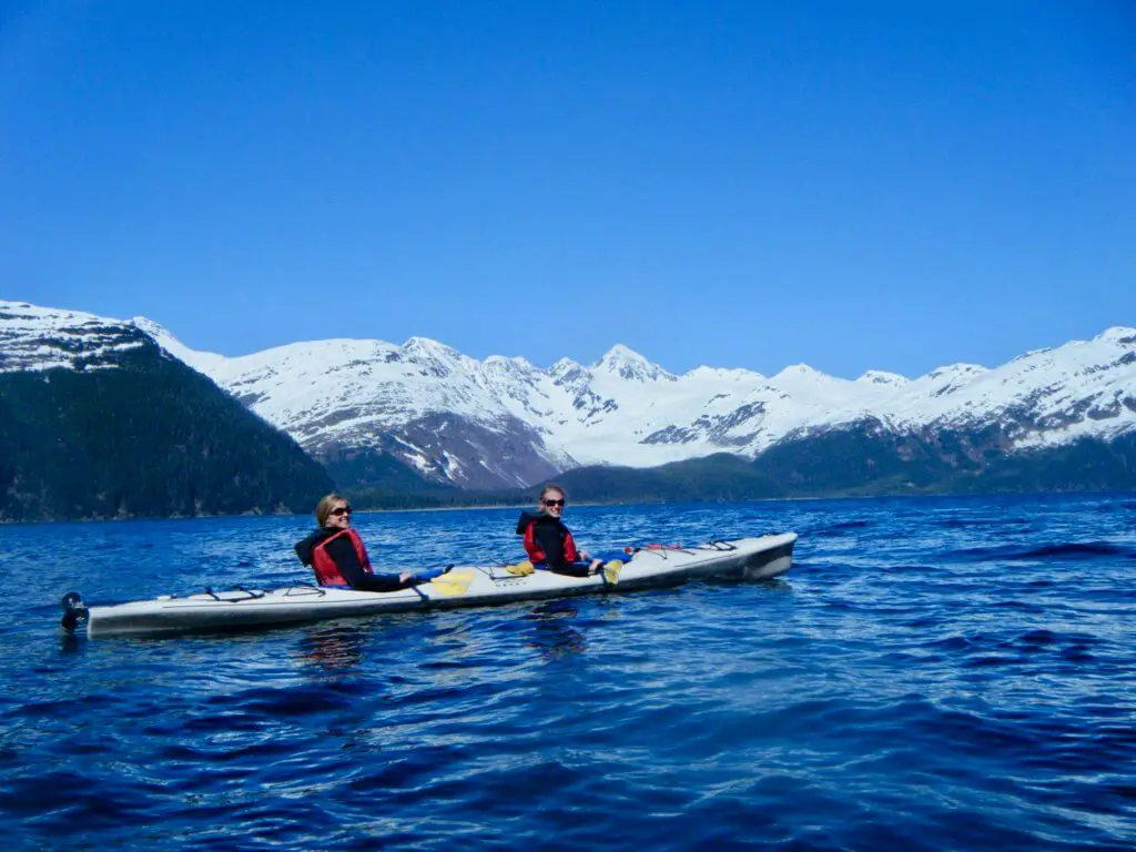 Gwen and a friend kayaking in front of snow-capped mountains. Sea kayaking is a great activity to check off your Alaska bucket list.