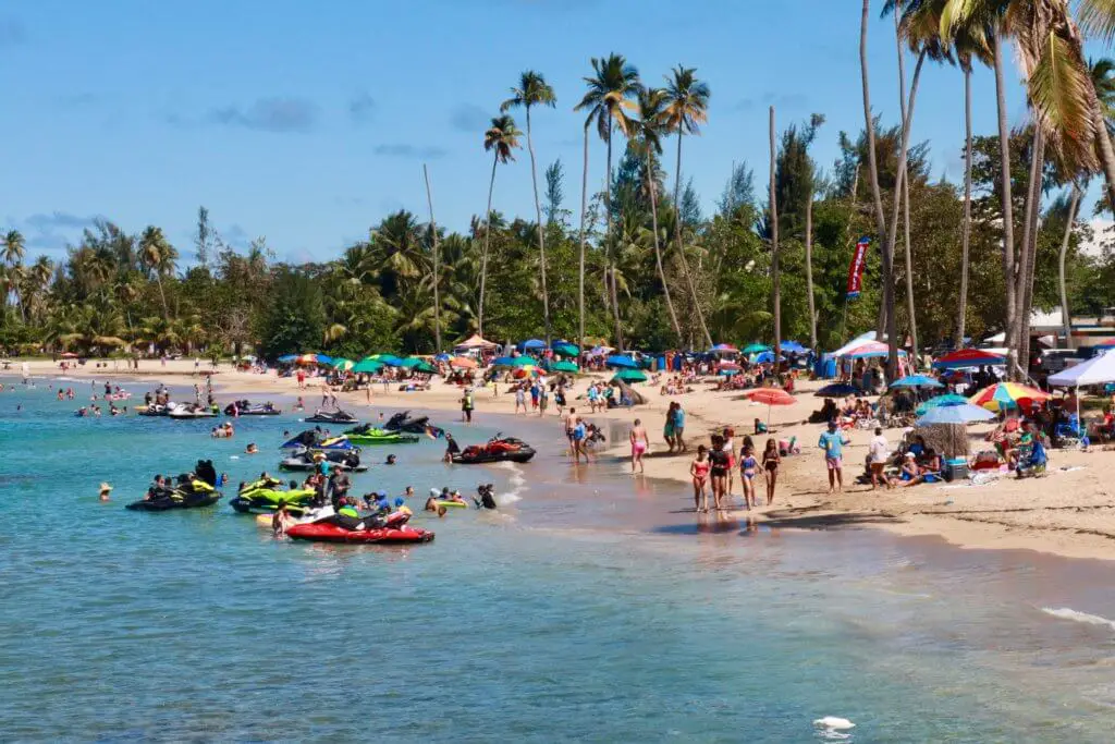 Beachgoers flanked by palm trees and umbrellas with jet skis in the teal water