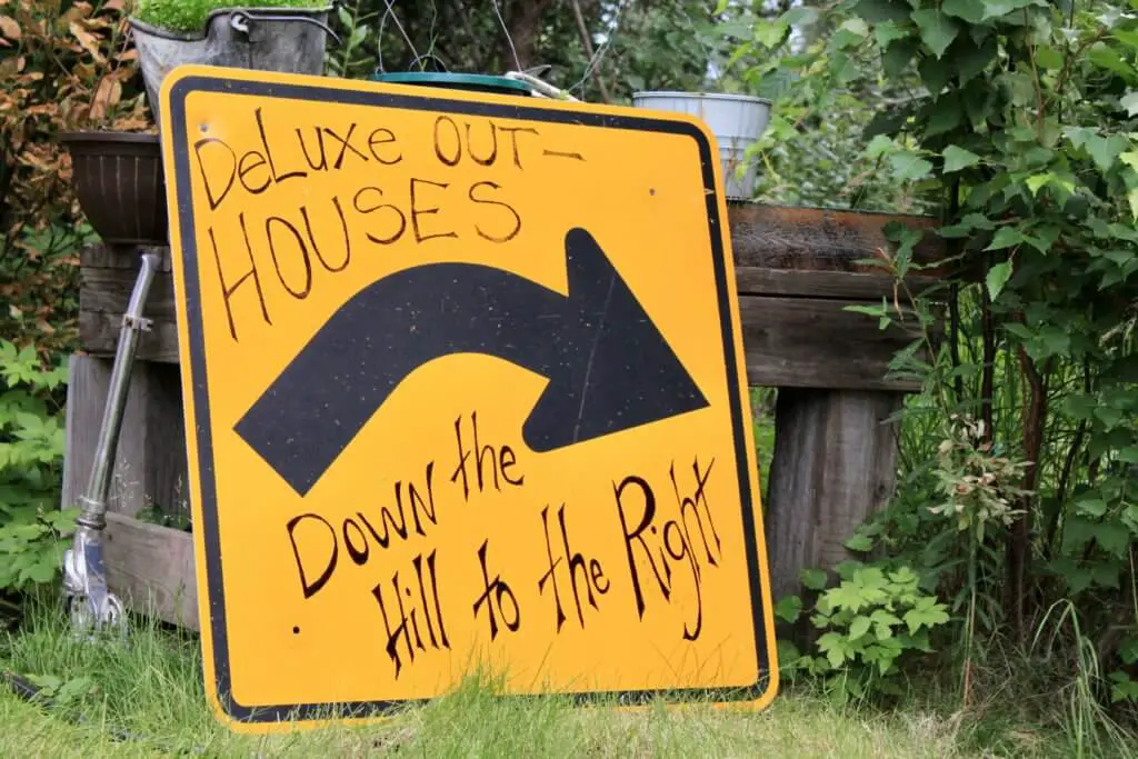 Road sign with hand-written letters saying "Deluxe outhouses down the hill to the right"