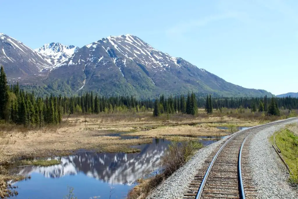 Railroad tracks cutting through forest and marshland in front of snow-capped mountains