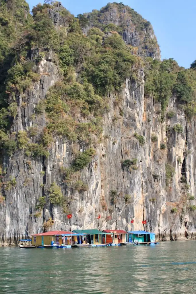 Small, colorful wooden shacks sitting on the water under a huge limestone cliff face